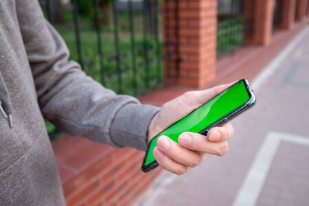Young man in gray sweatshirt holds a phone with green screen outsideの写真素材