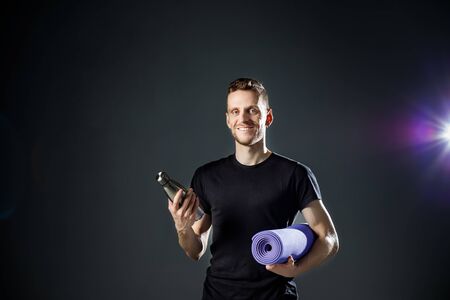 Young man on the black background is holding fitness bottle and fitness mat.の写真素材