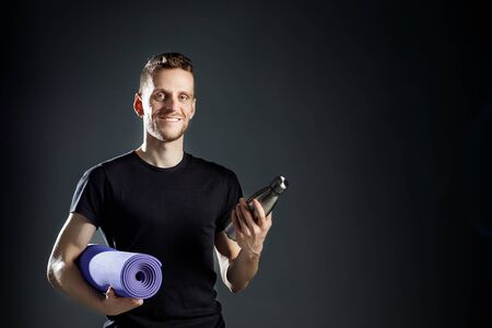 Young man on the black background is holding fitness bottle and fitness mat.の写真素材