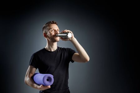 Young man on the black background is drinking water from fitness bottle, holding fitness mat.の写真素材