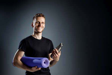 Young man on the black background is holding fitness bottle and fitness mat.