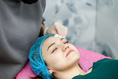 The beautician wipes the face with a napkin after procedures to the young girl lying on a couch