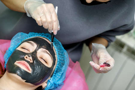The beautician applies a black mask for peeling the face of a young girl with a brush. A cosmetologist performs an exfoliation procedure on a young girl lying on a couch.の写真素材