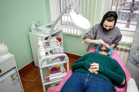 Kamyanets-Podilskyi, Ukraine - February 03, 2021: The beautician wipes the face with a napkin after procedures to the young girl lying on a couch.のeditorial素材