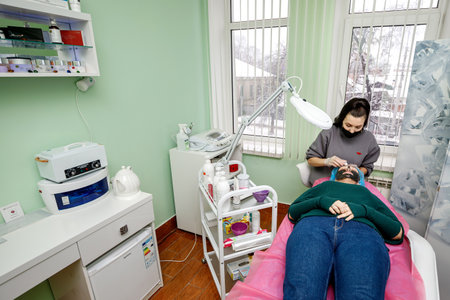 Kamyanets-Podilskyi, Ukraine - February 03, 2021: The beautician wipes the face with a napkin after procedures to the young girl lying on a couch.のeditorial素材