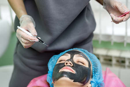 The beautician applies a black mask for peeling the face of a young girl with a brush. A cosmetologist performs an exfoliation procedure on a young girl lying on a couch.の写真素材