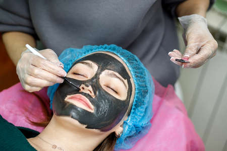 The beautician applies a black mask for peeling the face of a young girl with a brush. A cosmetologist performs an exfoliation procedure on a young girl lying on a couch.の写真素材