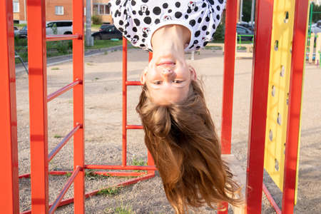 Happy girl child swinging upside down in the playground. Child plays on the horizontal bar and stairs.の写真素材