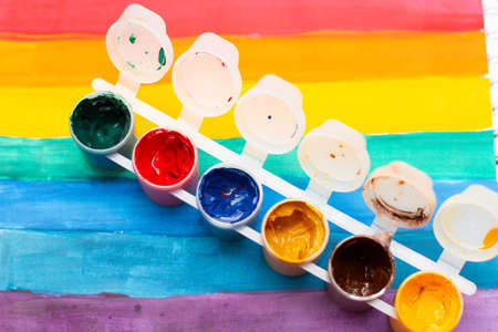 Jars of colorful gouache on a child's drawing of a rainbow in an album.