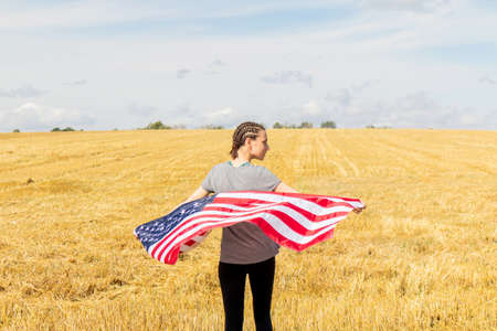 Young woman holding an American flag on the wheat field. USA flag waving on wind.