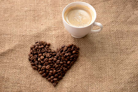 Heart made from coffee beans and Cup of fresh brewed coffee on brown cloth ragの写真素材