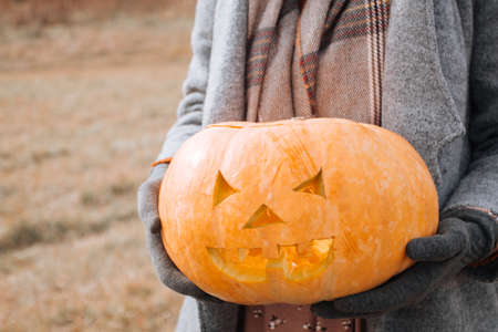 Woman in an autumn coat holding carved pumpkin against in autumn field.