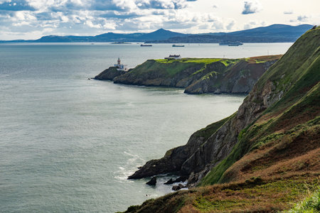 A natural landscape with a cliff overlooking the water, featuring a distant lighthouse against the sky. The horizon meets the sea in a picturesque view of nature. Howth, Irelandの写真素材