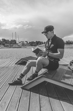 European man, a student in a black shirt and a cap reading a book in the city Park, near the riverの写真素材