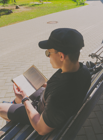 European man, a student in a black shirt and a cap reading a book in the city Park, near the riverの写真素材