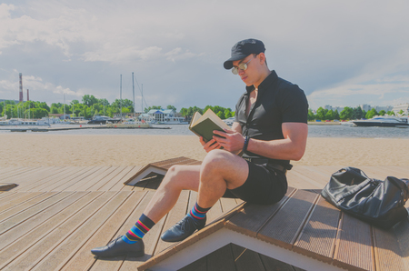 European man, a student in a black shirt and a cap reading a book in the city Park, near the riverの写真素材