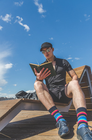 European man, a student in a black shirt and a cap reading a book in the city Park, near the riverの写真素材