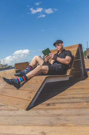European man, a student in a black shirt and a cap reading a book in the city Park, near the riverの写真素材