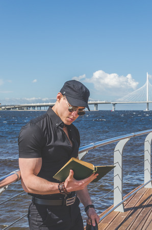European man, a student in a black shirt and a cap reading a book in the city Park, near the riverの写真素材