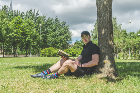 European man, a student in a black shirt and a cap reading a book in the city Park, near the riverの写真素材