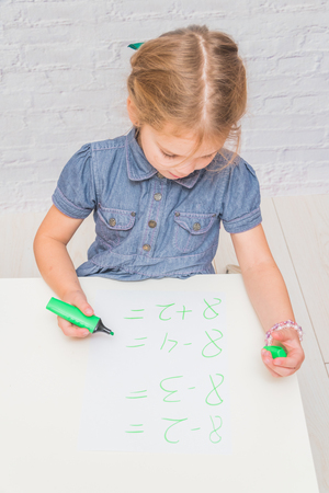 child, a girl at the table writes, draws on a piece of paper, against a white brick wallの写真素材