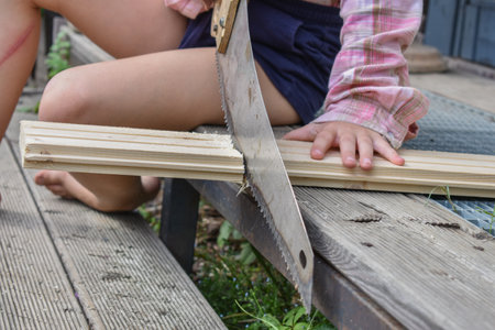 a child girl with a hacksaw saws a board outside photo without processingの写真素材