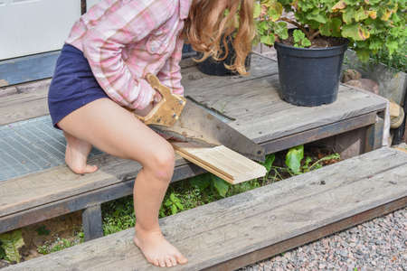 a child girl with a hacksaw saws a board outside photo without processingの写真素材