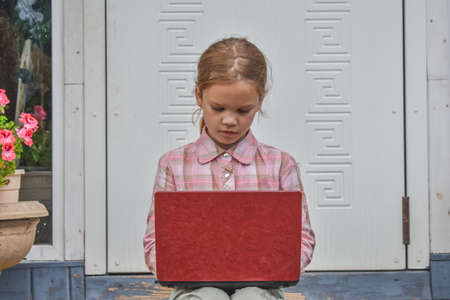 a girl child with a laptop is sitting on the porch of the house during a lesson on a remote photo without a filterの写真素材