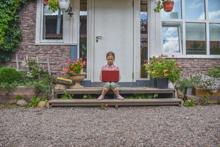 a girl child with a laptop is sitting on the porch of the house during a lesson on a remote photo without a filterの写真素材