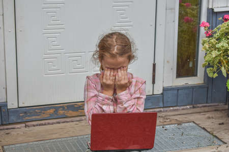 a girl child with a laptop is sitting on the porch of the house during a lesson on a remote photo without a filterの写真素材