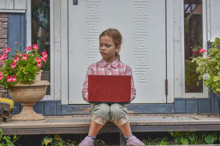 a girl child with a laptop is sitting on the porch of the house during a lesson on a remote photo without a filterの写真素材