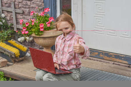 a girl child with a laptop is sitting on the porch of the house during a lesson on a remote photo without a filterの写真素材