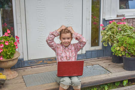a girl child with a laptop is sitting on the porch of the house during a lesson on a remote photo without a filterの写真素材
