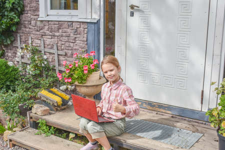 a girl child with a laptop is sitting on the porch of the house during a lesson on a remote photo without a filterの写真素材