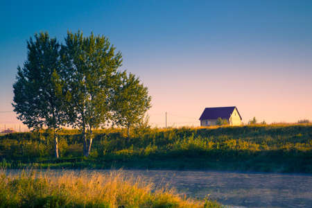 Lone house on banks of river on a summer morning with mistの写真素材