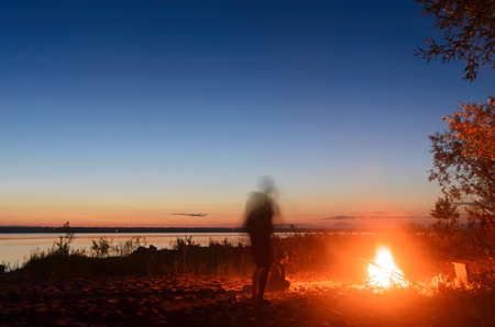 Silhouette of a man sitting by the fire and bowed his head, next to the blurred silhouette of the girl walking the beach at night beside a forest and river with a beautiful sky and sunset.の写真素材