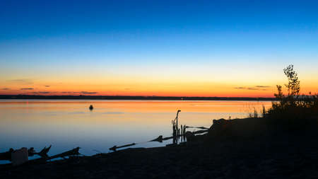 The dark silhouette of two lovers kissing in the water at night in the river on the horizon against a beautiful orange sunset sky at the shore with beach sand and the water tank at the camp.の写真素材