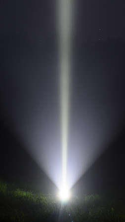 Bright beam led flashlight travel vertically from the lamp shines on the green grass to the blue of the sky with the stars over the trees on a dark night in the forest.の写真素材