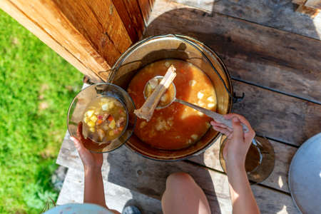 Girl chef holding spoon in bone and handing out soup in a transparent bowl soup of the boiler with a meal outdoors on the wooden floor in the forest.の写真素材