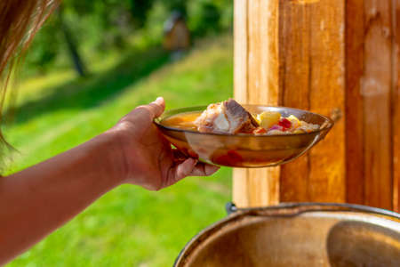 Hands girl cooks outdoors hold a plate of soup borscht with meat and vegetables on the background of the boiler with food in nature.の写真素材