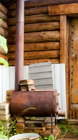 A small metal stove stands above the bricks in the grass in the Yakut Northern greenhouse for plants made of pine timber to heat the garden with wood in the cold.の写真素材