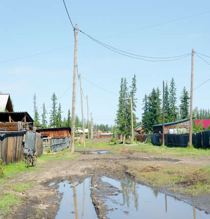A large puddle of mud on the residential street of the Northern Yakut village in the forest with a Bicycle at the fence and power lines on a bright day in the wild tundra.の写真素材