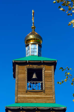Wooden chapel with a bell in the window and a Golden dome with a cross next to the leaves of yellow autumn birch.の写真素材