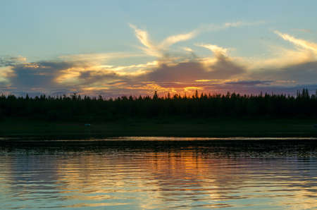Bright orange sunset behind the clouds of sun on the banks of the Northern river Viluy under the blue sky in the spruce forest and the silhouette of a car vacationer.の写真素材