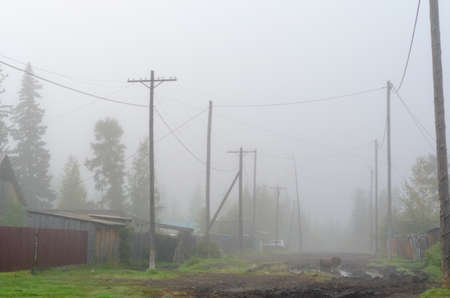 The street of the Northern Yakut village of ulus Suntar in the morning fog with puddles on the road and standing car behind the power line poles and wires.の写真素材