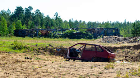 Abandoned burned the body of an old Russian car lying in the field on the background of abandoned after the fire farm buildings in the woods in the afternoon.の写真素材