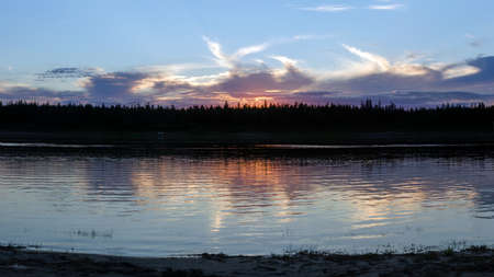 Bright orange sunset behind the clouds of sun on the banks of the Northern river Viluy under the blue sky in the spruce forest and silhouettes of cars and people resting in Yakutia.の写真素材