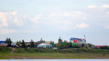 The road from the boats on the river Bank leads to the cliff with houses and silhouettes of cars going to the Church among the houses in the Northern village of ulus Suntar in Yakutia.の写真素材