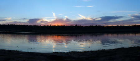 Panorama of a picturesque sunset on the banks of the Northern river Viluy under the sky with clouds and cliffs of spruce taiga in Yakutia with ripples from the wind.の写真素材