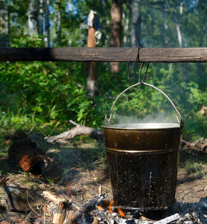 In a bucket hanging on a stick over a fire, soup is cooked in the wild Northern forest of Yakutia at a halt in the afternoon among the vegetation.の写真素材